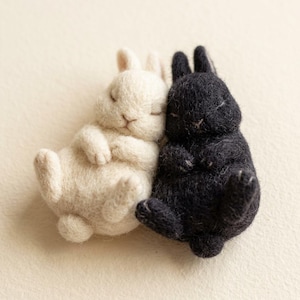 Pode incluir: Duas pequenas figuras de coelho em lã feltrada, uma de cor creme e outra preta, deitadas lado a lado. Ambos os coelhos têm olhos fechados, orelhas arredondadas e uma textura macia e felpuda, numa posição de repouso.
