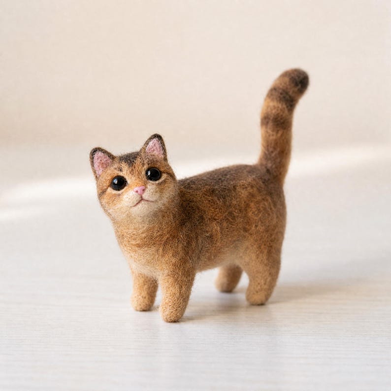 Pu&ograve; includere: Una statuetta di gatto in lana infeltrita con un mantello marrone e beige testurizzato, strisce marrone scuro sulla coda, grandi occhi neri e un piccolo naso rosa. Il gatto &egrave; in posizione quadrupedale con la coda alzata e la testa inclinata.