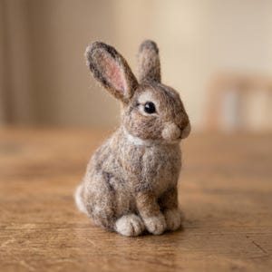 Pode incluir: Uma figura de coelho de feltro artesanal. O coelho é principalmente castanho com detalhes em castanho claro e branco. Possui orelhas longas com interior rosa, uma gola branca e está sentado em posição vertical. Um item decorativo charmoso.
