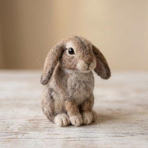 Könnte beinhalten: Eine aus Nadelfilz gefertigte Kaninchenfigur mit langen, herabhängenden Ohren und einem strukturierten Fell in Braun-, Beige- und Cremetönen. Die kleine Skulptur hat dunkle, runde Augen und ein flauschiges Aussehen, das einem Widderkaninchen ähnelt.