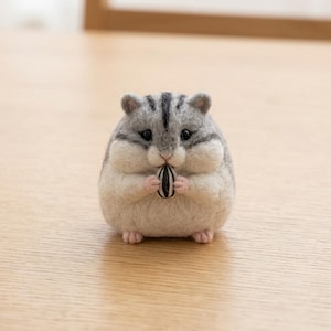 May include: A needle-felted hamster figurine with a round, plump body, featuring grey and white fur, small black eyes, and pink paws. The hamster is holding a single black and white striped sunflower seed between its front paws.