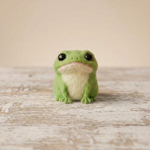 Puede incluir: Una figura de rana hecha con lana fieltrada con aguja en tonos verde y crema. La rana presenta un cuerpo redondeado, un vientre de color claro y dos ojos grandes de color negro brillante situados en la parte superior de la cabeza. Es suave.