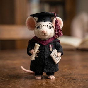 Puede incluir: Figura de ratón artesanal vestida con toga de graduación, birrete y gafas redondas. El ratón sostiene un diploma y un pequeño libro que dice "Class of '24". La toga es negra con cuello y borla burdeos.