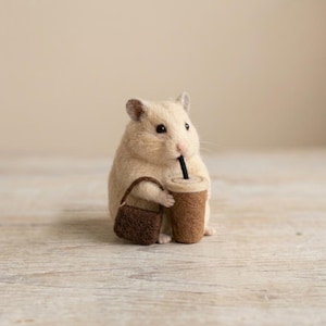 Könnte beinhalten: Eine nadelgefilzte, beige Hamsterfigur, die einen braunen Filzbecher mit schwarzem Strohhalm und eine kleine, dunkelbraune Filztasche hält. Der Hamster hat schwarze Augen, eine rosa Nase und abgerundete Ohren mit einer strukturierten Oberfläche.