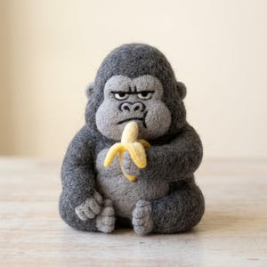Könnte beinhalten: Eine nadelgefilzte Gorillafigur aus dunkelgrauer und hellgrauer Wolle. Der Gorilla hat einen runden Körper, einen strengen Gesichtsausdruck und hält eine kleine, gelbe, teilweise geschälte Banane in seinen Händen. Die Textur ist flauschig.
