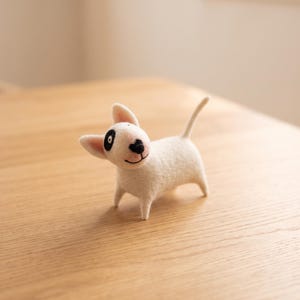 May include: A small, white needle-felted bull terrier figurine with a black patch over one eye, a black nose, and a stitched mouth. The dog stands on four short legs with its tail pointed upward, featuring a textured, fuzzy wool surface.