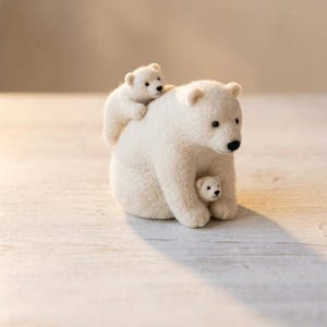 May include: A needle-felted sculpture of a large white polar bear with two smaller polar bear cubs. One cub is positioned on the back of the adult bear, while the second cub is nestled between the front paws of the adult bear. All three bears have black eyes and noses.