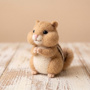 May include: A needle-felted chipmunk figurine with a tan and cream-coloured body, dark brown stripes along its back, a small pink nose, and black bead eyes. The small animal is depicted in a standing position with its front paws held together.