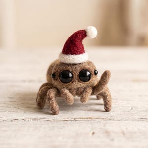 Op de afbeelding: Een naaldgevilte bruine spinnenfiguur met twee grote zwarte ogen en een kleine rood-witte kerstmuts op het hoofd. De spin heeft meerdere getextureerde poten en een pluizig, rond lichaam, vervaardigd uit bruine wolvezels in een zachte tint.