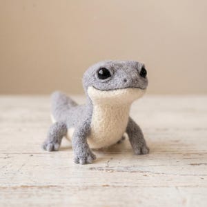 May include: A needle-felted lizard figurine crafted from soft wool fibres. The creature features a textured grey back and head with a smooth, cream-coloured underbelly. It has two large, glossy black bead eyes and four small, detailed feet.