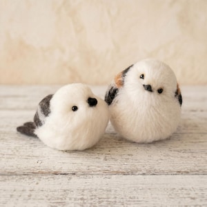 Puede incluir: Dos figuras de pájaros redondas de lana fieltrada con cuerpos blancos esponjosos, pequeños picos negros y ojos oscuros. Un pájaro presenta marcas grises y blancas en las alas, mientras que el otro muestra acentos grises, blancos y marrones claros.