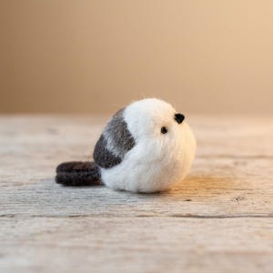 以下が含まれることがあります： 手作りのフェルトの鳥の置物。鳥は白い体、背中の灰色のパッチ、濃い灰色の尾を持っています。小さな黒い目と黒いくちばしがあります。鳥は木製の表面に座っています。
