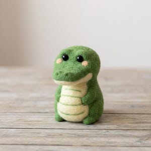 May include: A small, needle-felted crocodile figurine crafted from green and cream-coloured wool. The creature features a rounded head, two black bead eyes, small pink cheek patches, and a textured belly with horizontal lines across the front.