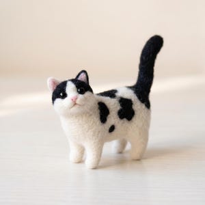 Pu&ograve; includere: Una statuetta di gatto in lana infeltrita con corpo bianco e macchie nere sulla testa, sulla schiena e sulla coda. Il gatto ha piccole orecchie rosa, un minuscolo naso rosa e occhi a perla scuri e lucidi. La coda &egrave; tenuta in posizione verticale.