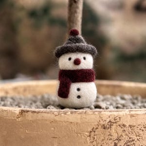May include: A handcrafted snowman figurine made of white felt, wearing a dark gray hat and a burgundy scarf. The snowman has a red nose and black button eyes and a button mouth. The figurine is a festive decoration.
