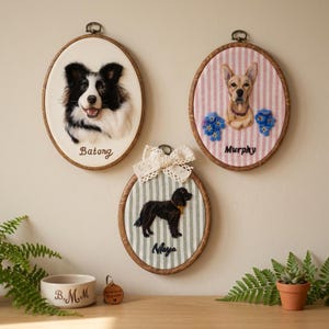 以下が含まれることがあります： ニードルフェルトで描かれた犬の肖像画が入った3つの楕円形の木製刺繍枠。1つはバトンという名前の白黒の犬、もう1つは青い花を添えたマーフィーという名前の茶色の犬、3つ目はレースのリボンが付いたマヤという名前の黒い犬が描かれています。