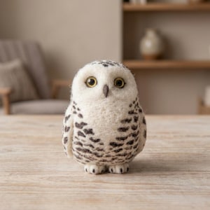 May include: A needle-felted snowy owl figurine featuring a round, off-white body with dark brown, speckled markings across the wings and chest. The owl has large, round, yellow eyes with black pupils and a small, dark, pointed beak.