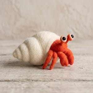 May include: A needle-felted hermit crab figurine featuring a bright orange body with two black-eyed stalks and a textured, off-white spiral seashell. The soft, fuzzy creature is crafted with visible wool fibers and displays a detailed, rounded shell.