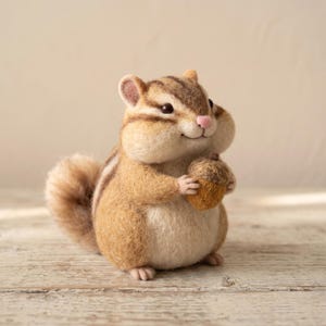 May include: A needle-felted chipmunk figurine with a round body, brown and cream fur, dark stripes on its head and back, and a small pink nose. The chipmunk is holding a textured brown acorn in its front paws and has a fluffy, curved tail.