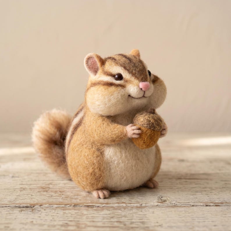 May include: A needle-felted chipmunk figurine with a round body, brown and cream fur, dark stripes on its head and back, and a small pink nose. The chipmunk is holding a textured brown acorn in its front paws and has a fluffy, curved tail.