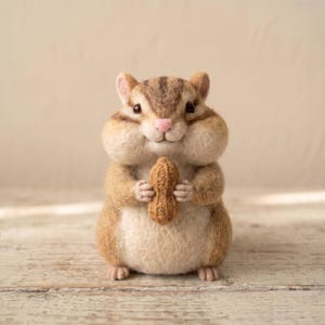 May include: A needle-felted chipmunk figurine with brown and cream fur, dark stripes on its head, and a small pink nose. The chipmunk has full cheeks and is holding a textured brown peanut in its front paws, standing in an upright position.