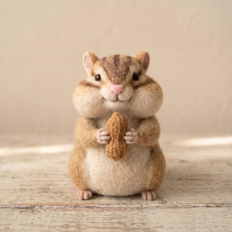 May include: A needle-felted chipmunk figurine with brown and cream fur, dark stripes on its head, and a small pink nose. The chipmunk has full cheeks and is holding a textured brown peanut in its front paws, standing in an upright position.