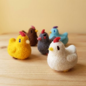 Peut inclure: Six figurines de poulets en feutre de différentes couleurs, dont jaune, blanc, gris, marron et bleu. Chaque poulet a une crête rouge et un bec jaune. Les figurines sont disposées sur une surface en bois clair.