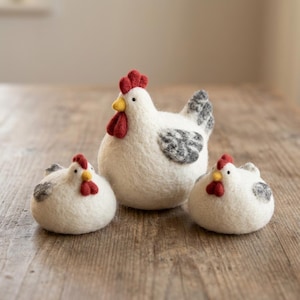 May include: Three needle-felted wool chicken figurines in varying sizes. Each chicken is white with a red comb and wattle, a yellow beak, and grey-speckled wings. The figures have rounded bodies and small black bead eyes, arranged in a group.