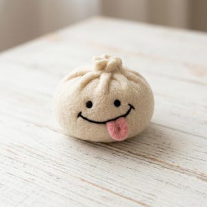 Könnte beinhalten: Ein rundes, cremefarbenes Knödel-Ornament aus gefilzter Wolle mit einem handgestickten schwarzen Gesicht, zwei runden Augen und einem breiten, geschwungenen Lächeln. Eine kleine, rosa gefilzte Zunge ragt aus dem Mund und oben ist ein Knoten.