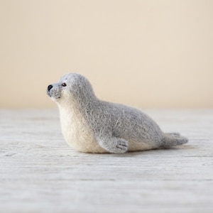 Op de afbeelding: Een naaldgevilte zeehondfiguur met een textuurrijke grijze rug en een crèmekleurige onderkant. Het kleine dierenbeeldje heeft een ronde kop, een donkere neus en donkere ogen, en het lichaam bevindt zich in een rusthouding op een vlak oppervlak.
