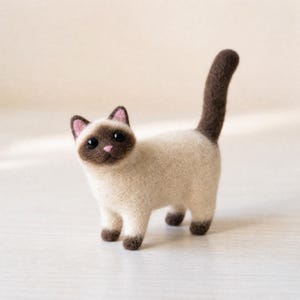 Pu&ograve; includere: Una statuetta di gatto siamese in lana infeltrita con corpo color crema e orecchie, muso, coda e zampe marrone scuro. Il gatto ha occhi a perla neri, un piccolo naso rosa e l'interno delle orecchie rosa. La figura &egrave; in piedi con la coda alzata.