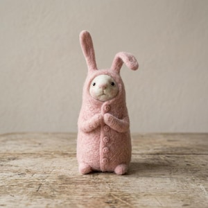Puede incluir: Una figura de conejo de fieltro rosa hecha a mano que lleva un mono con capucha con botones. El conejo tiene orejas largas y está de pie sobre una superficie de madera. Las manos del conejo están juntas.