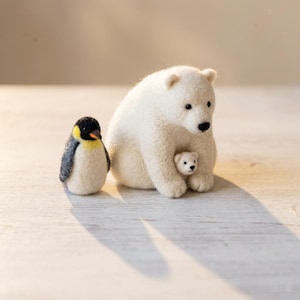 Op de afbeelding: Een naaldgevilte witte ijsbeer die zit met een kleine ijsbeerwelp tegen zijn zij, vergezeld door een staande pinguïn met een zwarte kop, gele nekvlek en grijze vleugels, alles gemaakt van zachte, getextureerde wolvezels.