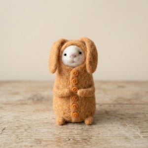 Pode incluir: Figura de coelho de feltro artesanal, vestindo um casaco laranja com capuz e botões. O coelho tem um rosto branco, nariz rosa e olhos pretos. A figura está sobre uma superfície de madeira.