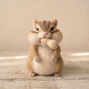 May include: A needle-felted chipmunk figurine with brown and cream fur, dark stripes on its head, and a small pink nose. The chipmunk is holding a textured, light brown peanut in its mouth, with its front paws resting against its round belly.
