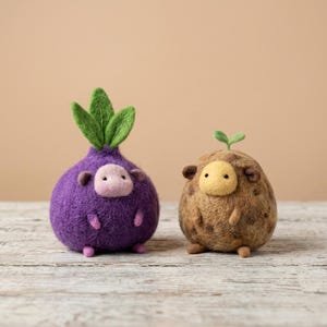 May include: Two needle-felted figures. The left figure is a round, purple creature with a light pink face, small ears, and three green leaves on top. The right figure is a round, brown creature with a yellow face, small ears, and one green sprout.