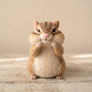 May include: A needle-felted chipmunk figurine with a rounded body, textured tan and brown fur, dark brown stripes on its head, small black eyes, and a tiny pink nose. The chipmunk is standing upright with its small front paws held near its cheeks.
