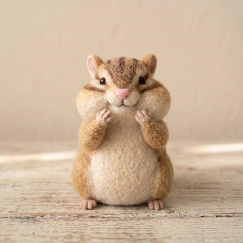 May include: A needle-felted chipmunk figurine with a rounded body, textured tan and brown fur, dark brown stripes on its head, small black eyes, and a tiny pink nose. The chipmunk is standing upright with its small front paws held near its cheeks.