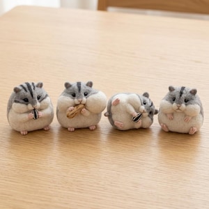 Könnte beinhalten: Vier gefilzte Hamsterfiguren in verschiedenen Posen. Jeder Hamster hat ein grau-weißes Fell mit einem dunklen Streifen auf dem Rücken. Einer hält einen Sonnenblumenkern, einer eine Erdnuss, einer liegt auf dem Rücken mit einem Kern und einer sitzt.