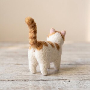 Pu&ograve; includere: Una statuetta di gatto in lana infeltrita ad ago, con corpo bianco, macchie arancioni sul dorso e una lunga coda arancione dritta. Il gatto &egrave; visto da dietro, mostrando le piccole orecchie rosa e la superficie in lana, in piedi su quattro zampe.