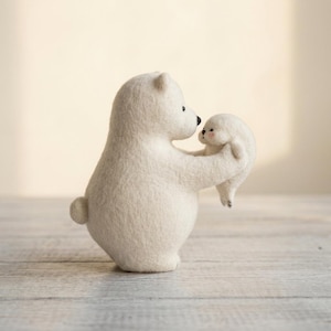 Może przedstawiać: Filcowana figurka niedźwiedzia polarnego w kolorze kremowym, trzymająca małą białą fokę. Niedźwiedź ma zaokrąglony korpus, mały ogon i czarne oczy z koralików. Foka ma maleńkie czarne oczy, czarny nos i delikatny różowy rumieniec na policzkach.