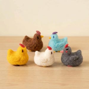 Peut inclure: Six figurines de poulets en feutre de différentes couleurs : jaune, marron, blanc, bleu et gris. Chaque poulet a une crête rouge ou violette et un bec jaune. Les figurines sont disposées sur une surface en bois brun clair.