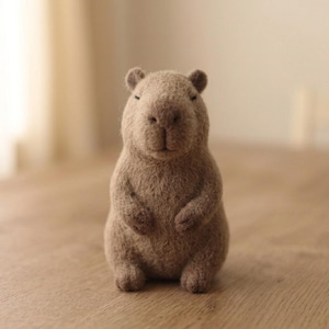 May include: A needle-felted capybara figurine crafted from light brown wool fibres. The small animal sculpture features a rounded body, short limbs, small ears, and a textured surface. The eyes are represented by two simple, dark horizontal stitches.