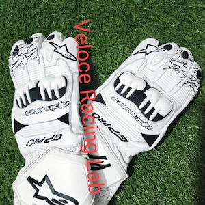 Könnte beinhalten: Weiße und schwarze Motorradhandschuhe mit dem Text "GP Pro" und "Alpinestars". Die Handschuhe haben eine Schutzpolsterung und ein Sternlogo. Die Handschuhe liegen auf einer grünen Oberfläche.