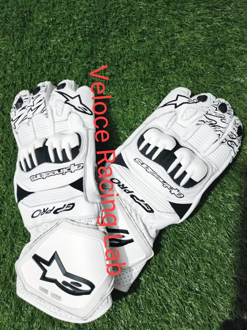 Op de afbeelding: Witte en zwarte motorhandschoenen met de tekst "GP Pro" en "Alpinestars". De handschoenen hebben beschermende vulling en een sterlogo. De handschoenen liggen op een groene ondergrond.