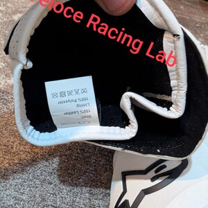Op de afbeelding: Witte en zwarte racehandschoen met de tekst "Veloce Racing Lab". De binnenkant is zwart met een label met 100% polyester voering en 100% leren schaal. De handschoen heeft een stervormig logo.