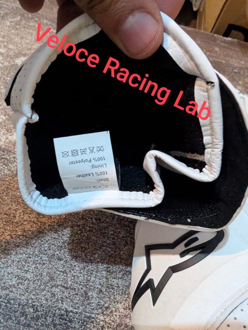Op de afbeelding: Witte en zwarte racehandschoen met de tekst "Veloce Racing Lab". De binnenkant is zwart met een label met 100% polyester voering en 100% leren schaal. De handschoen heeft een stervormig logo.