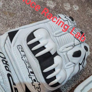 Op de afbeelding: Witte en zwarte motorhandschoenen met de tekst "GP Pro" en "Alpinestars". De handschoenen hebben beschermende elementen op de knokkels en vingers. De tekst "Veloce Racing Lab" is ook zichtbaar.
