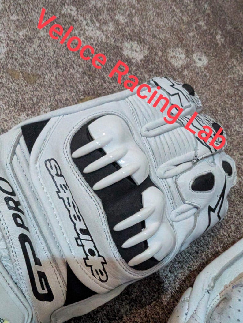Op de afbeelding: Witte en zwarte motorhandschoenen met de tekst "GP Pro" en "Alpinestars". De handschoenen hebben beschermende elementen op de knokkels en vingers. De tekst "Veloce Racing Lab" is ook zichtbaar.