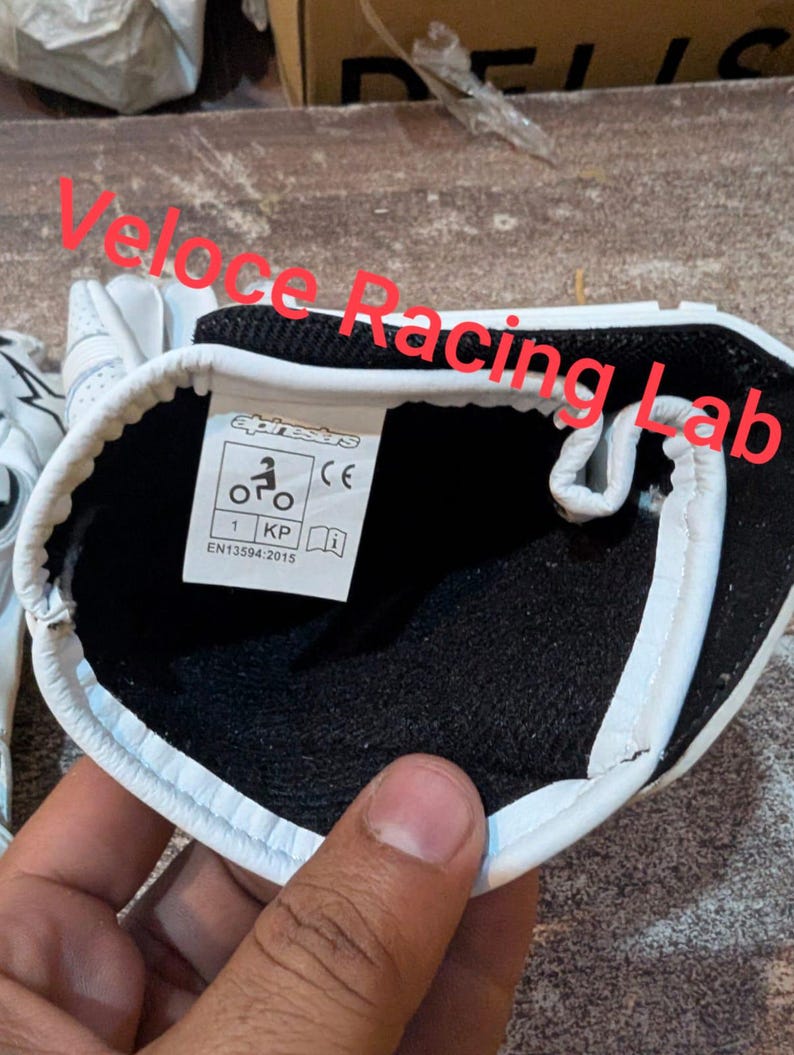Op de afbeelding: Witte motorhandschoenen met zwarte voering en witte rand. De handschoenen hebben een label met de tekst "alpinestars" en symbolen. Ontworpen voor motorraces, ze zijn gemaakt van duurzaam materiaal.